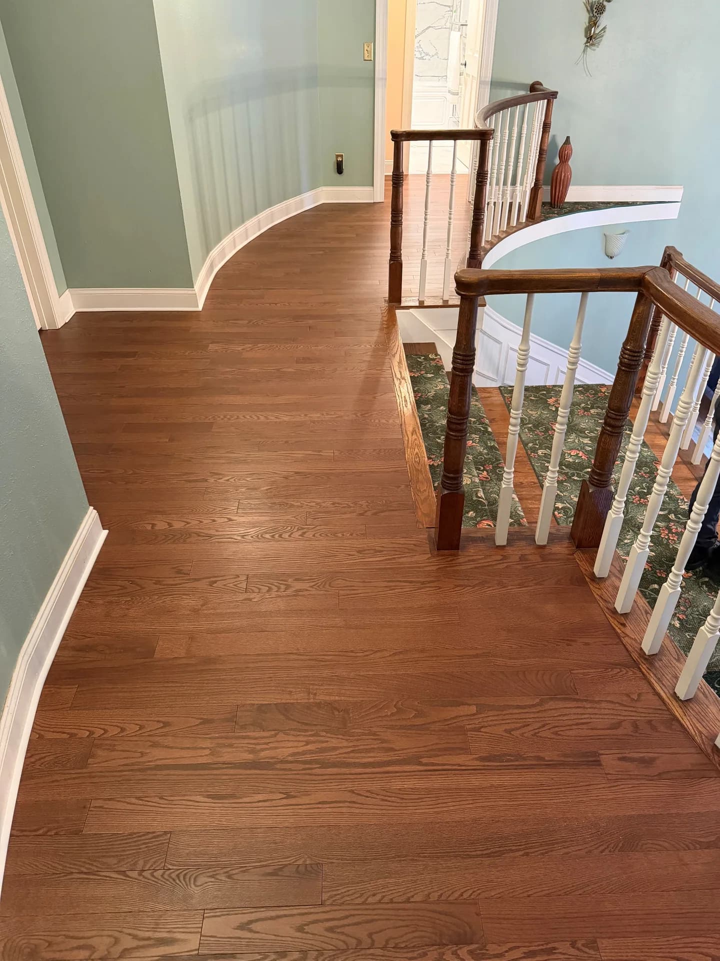 Completed hardwood hallway and landing in Southington, with a curved wall, white baseboards, and an existing railing beside carpeted stairs.