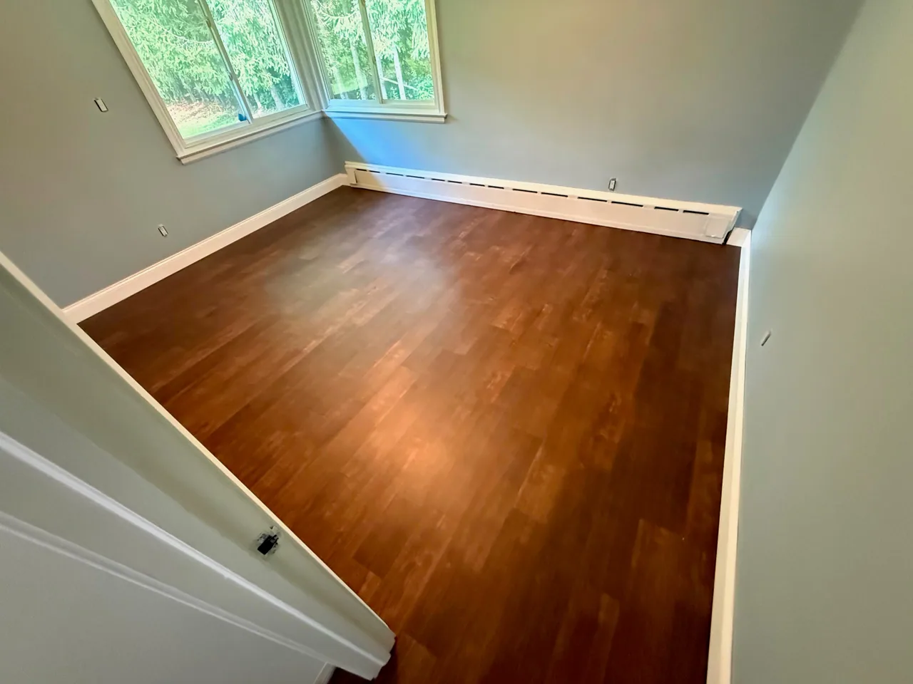 Brown plank flooring in an empty room with a bay window and baseboard heating.