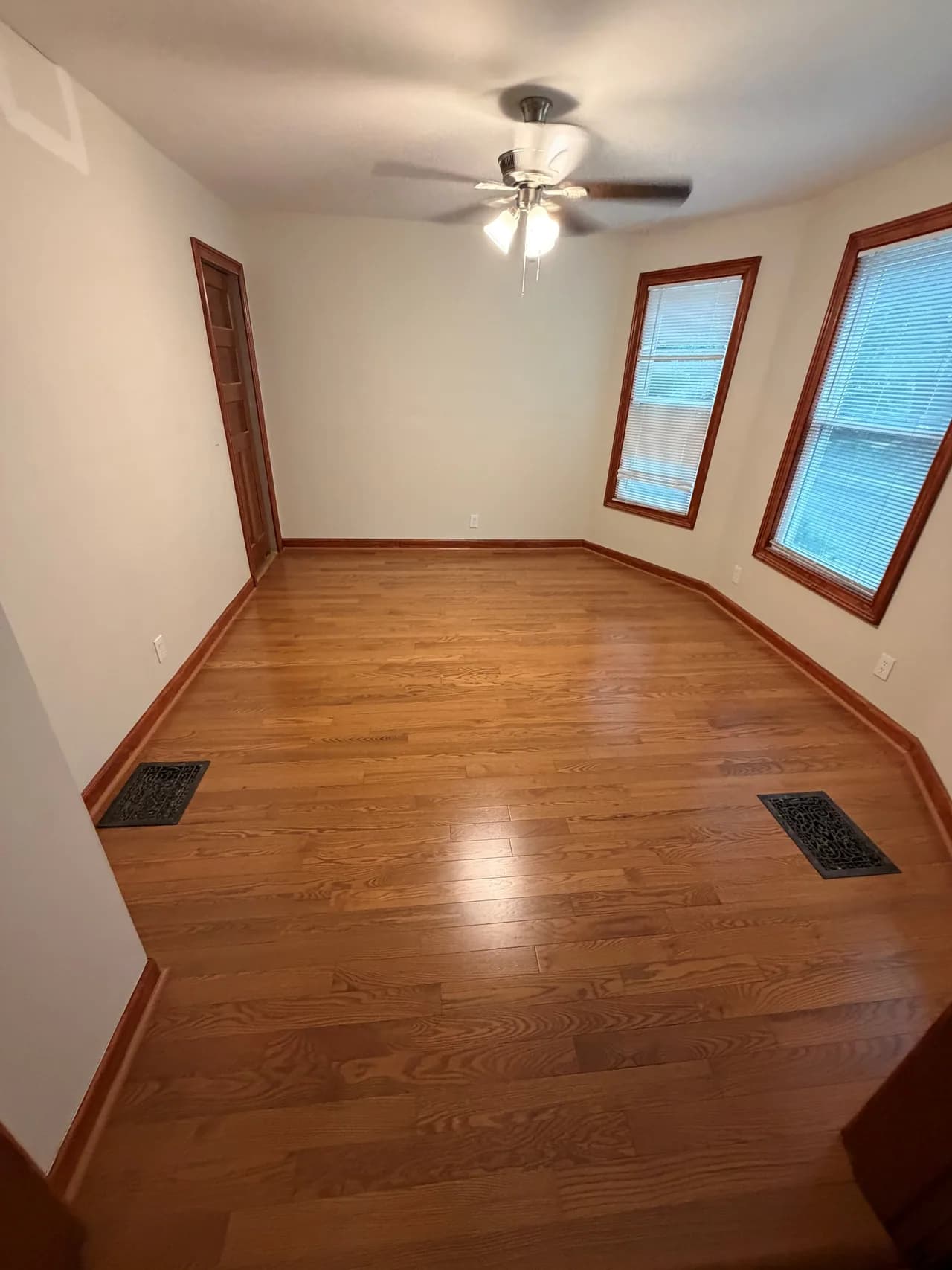 Wood strip flooring around two floor vents in an empty room with wood window trim.