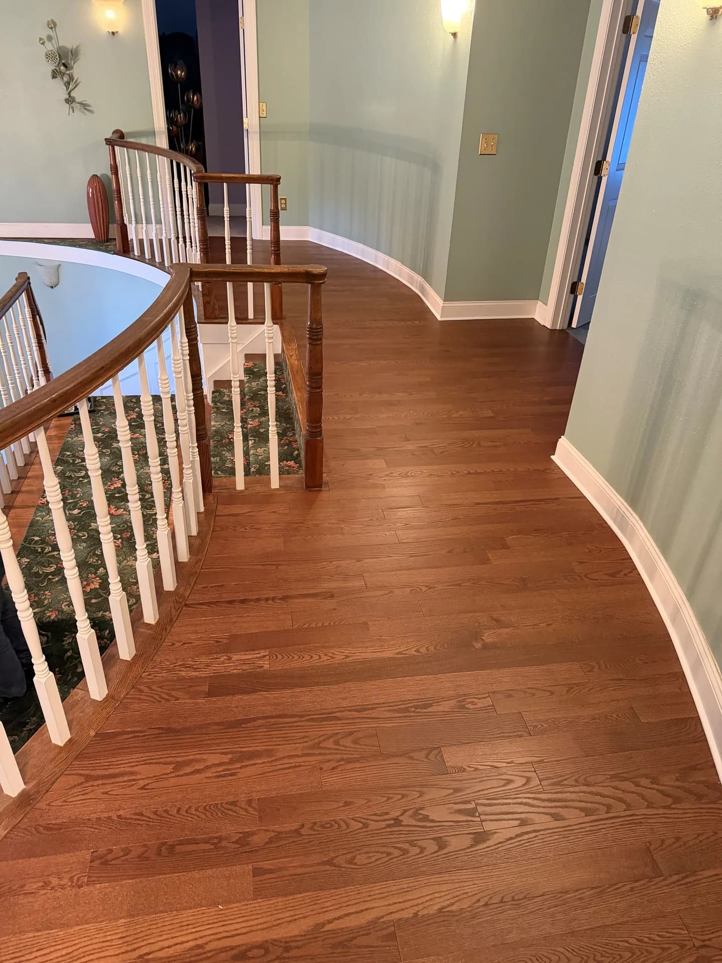 Brown hardwood flooring along a curved hallway wall and beside an existing stair railing in Southington, with white baseboards and adjoining doorways.